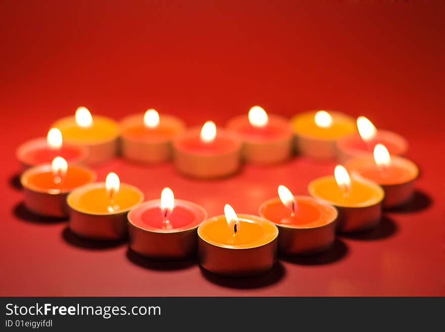 Small light candles, in heart form on a red background. Focus on the first, front candle. Small light candles, in heart form on a red background. Focus on the first, front candle.