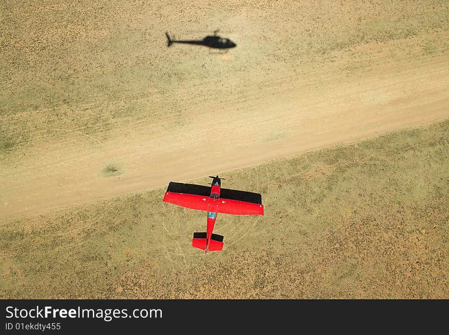 Aerial photograph of a red Cessna airplane next to a dirt airstrip with shadow of a helicopter