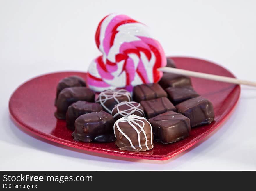 A variety of chocolates with a heart shaped lollipop on a red metallic heart plate isolated on white. A variety of chocolates with a heart shaped lollipop on a red metallic heart plate isolated on white