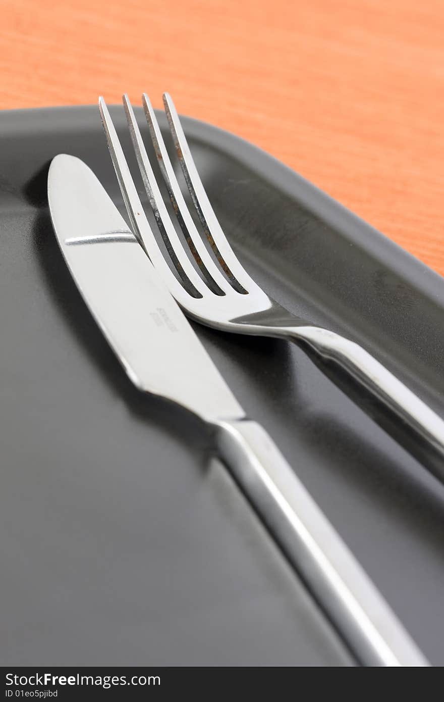 Detail of a knife and fork at a black dinner plate with a orange background. Detail of a knife and fork at a black dinner plate with a orange background