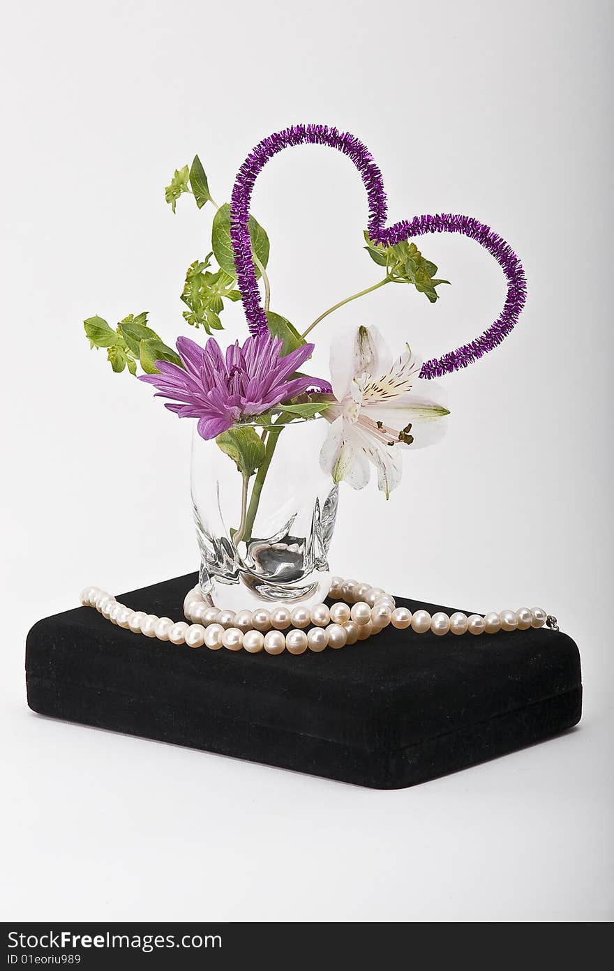 Valentine's day still life: a flower arrangement in a glass with a purple heart and a string of pearls all on a black jewelry box with a white background. Valentine's day still life: a flower arrangement in a glass with a purple heart and a string of pearls all on a black jewelry box with a white background