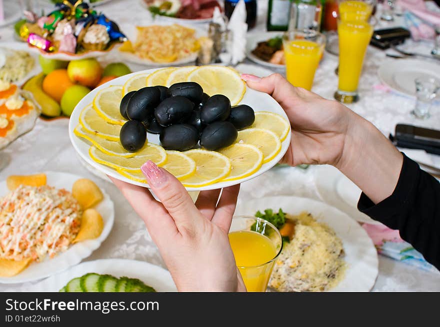 Black olives and lemons on a plate in female hands over a celebratory table. Black olives and lemons on a plate in female hands over a celebratory table.