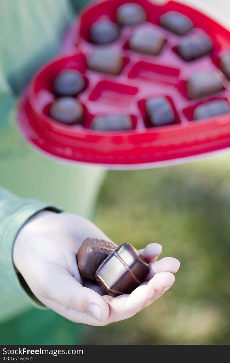 A young child is handing out two pieces of valentine chocolate from a heart shaped box while giving the chocolate in his hands