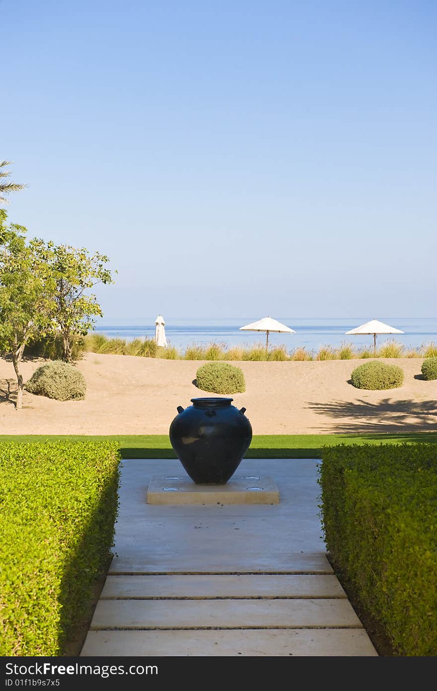 The beach exit from a lovely garden with a ceramic pot on display. Beach umbrellas are visible on the seaside. The beach exit from a lovely garden with a ceramic pot on display. Beach umbrellas are visible on the seaside.