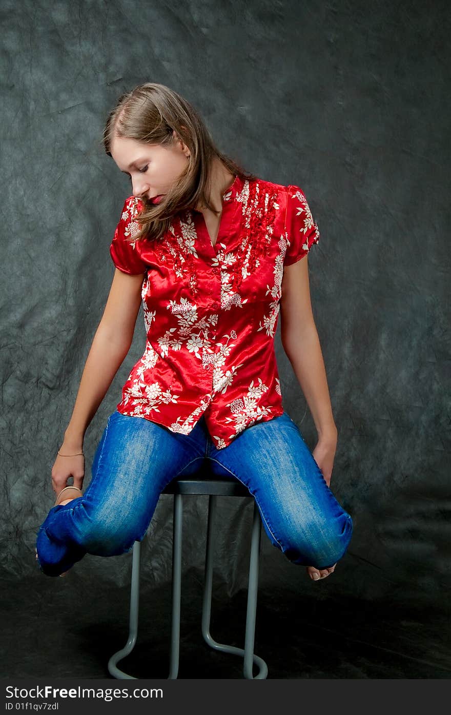 Young dark haired girl sitting on chair with legs lifted up and hands touching hips looking down thinking isolated on black background