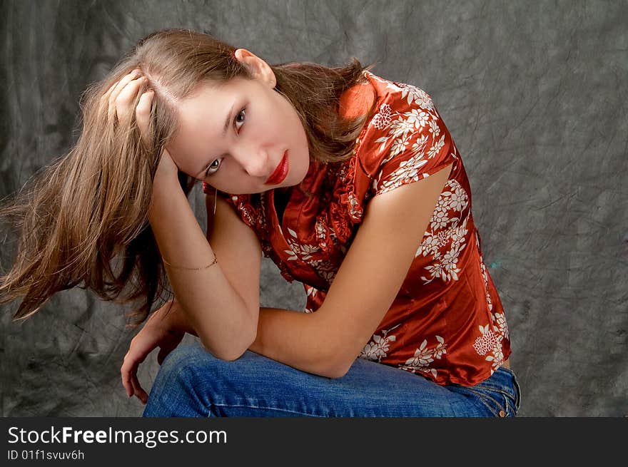 Pretty young girl sitting turned with hand lifted and touching hair legs crossed appear looking forward thinking isolated on gray background. Pretty young girl sitting turned with hand lifted and touching hair legs crossed appear looking forward thinking isolated on gray background