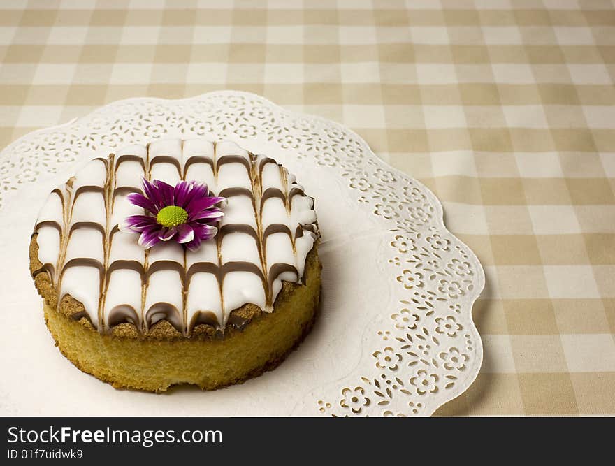 Baked fancy cake with a purple flower on a table with soft beige square cloth.