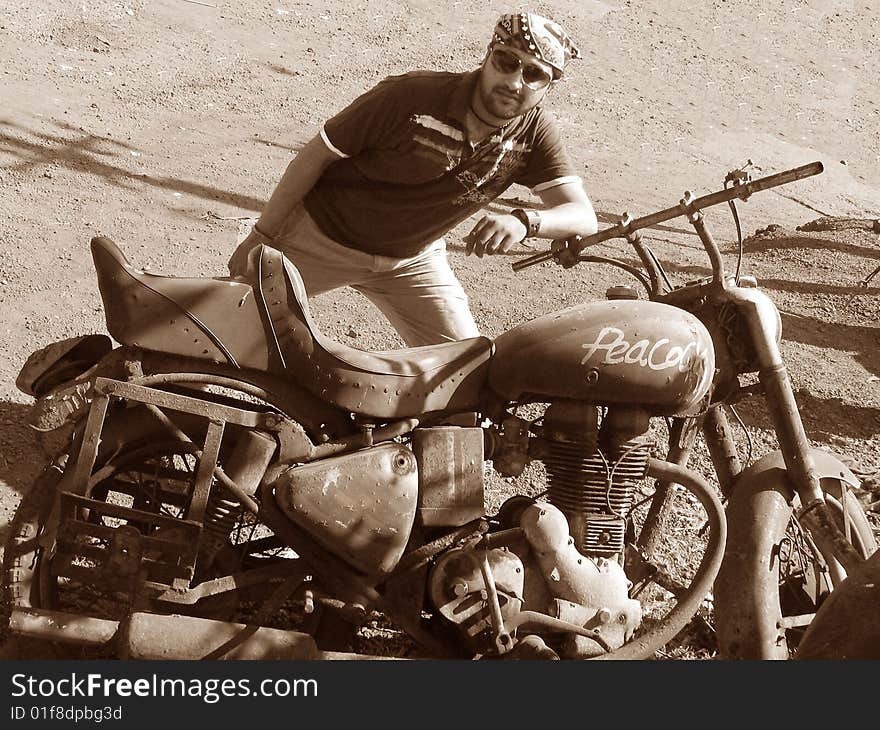 A young handsome guy posing besides a rusty old bike named peacock. A young handsome guy posing besides a rusty old bike named peacock