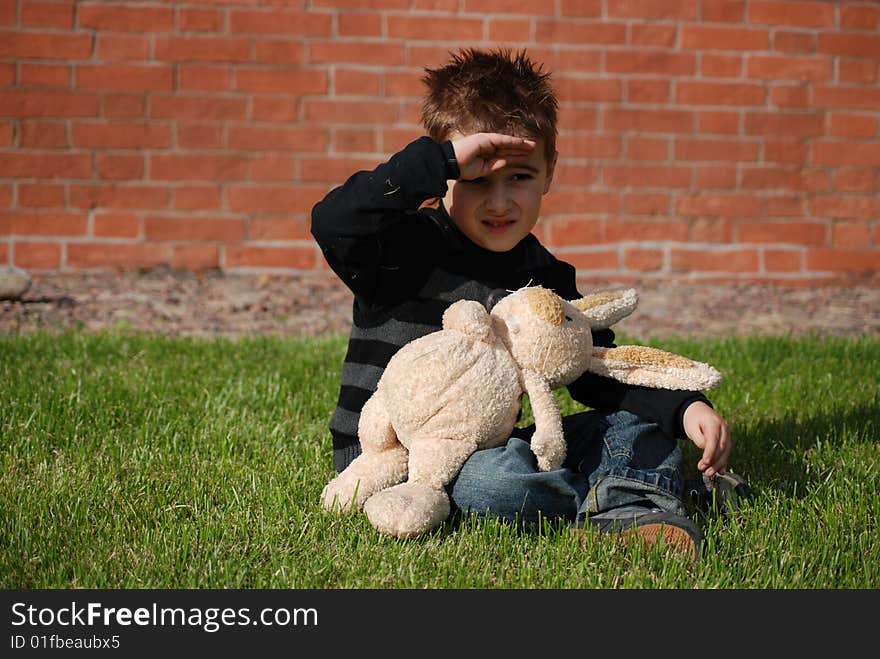 Little boy with a toy hare in hands, sitting on a green grass on a lawn on a background a brick wall. Little boy with a toy hare in hands, sitting on a green grass on a lawn on a background a brick wall