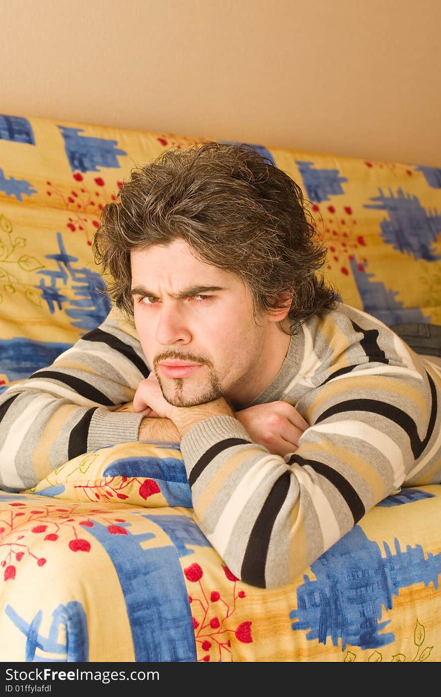 Young smiley dark haired caucasian man in striped sweater lying on country style sofa and frowning. Young smiley dark haired caucasian man in striped sweater lying on country style sofa and frowning