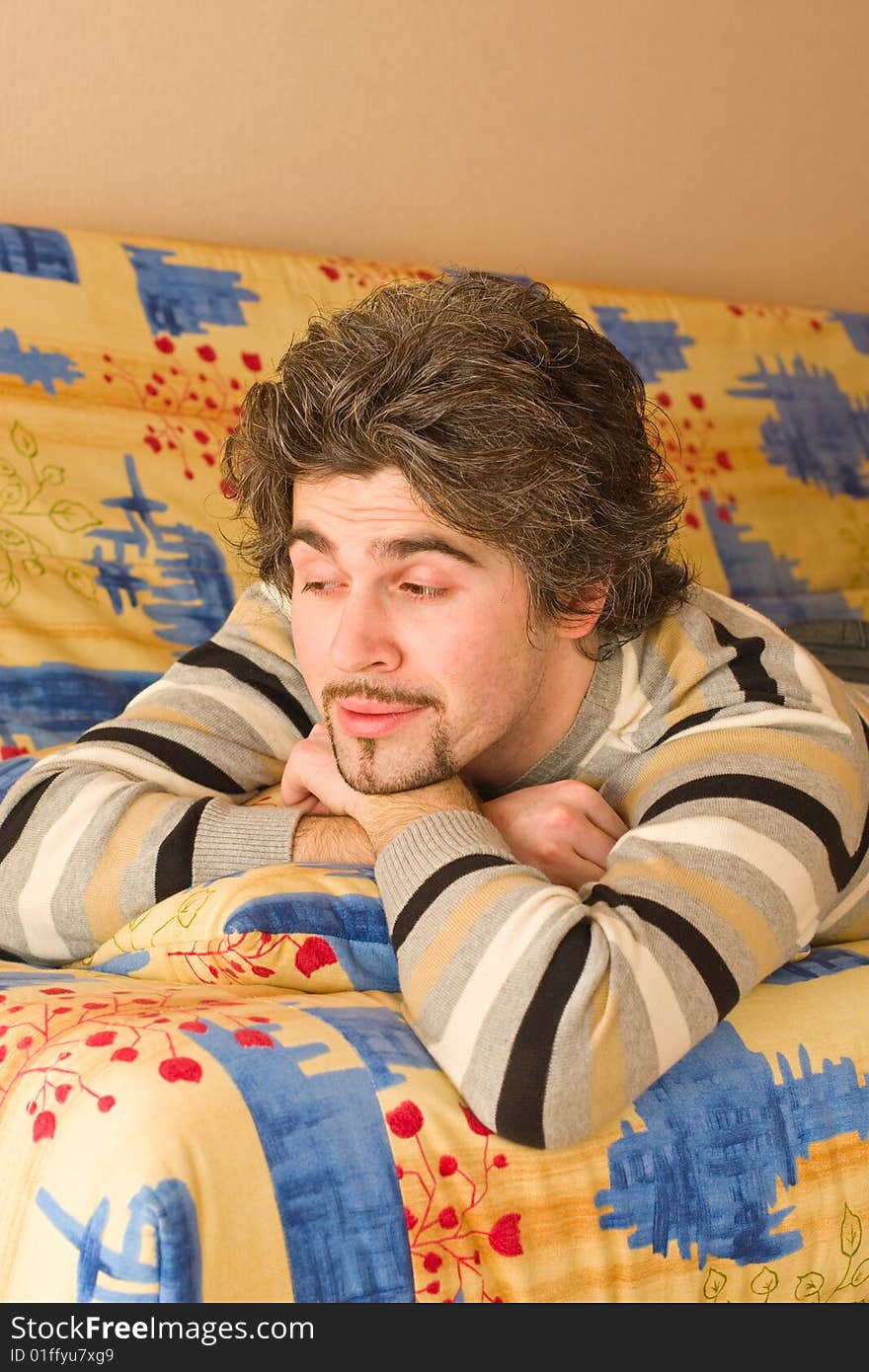 Young smiley dark haired caucasian man in striped sweater lying on country style sofa. Young smiley dark haired caucasian man in striped sweater lying on country style sofa