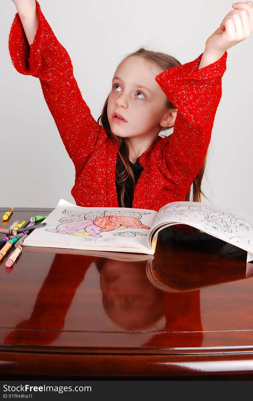 Little girl in a red sweater sitting on the table and coloring an book with crayons. Her face mirroring in the table and the arms are raised. Little girl in a red sweater sitting on the table and coloring an book with crayons. Her face mirroring in the table and the arms are raised.