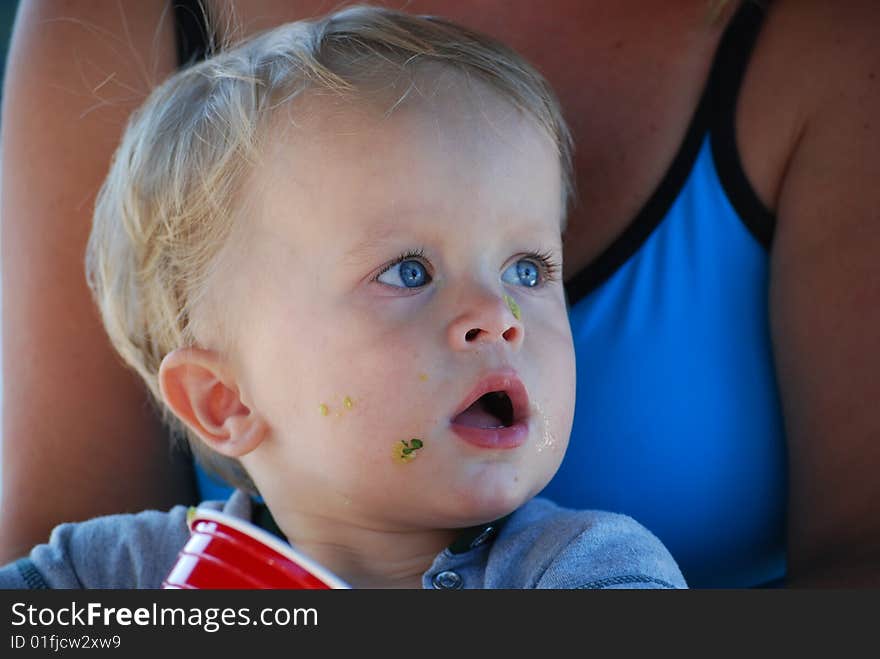 A little boy has guacamole out of a cup while sitting on his mother's lap. A little boy has guacamole out of a cup while sitting on his mother's lap