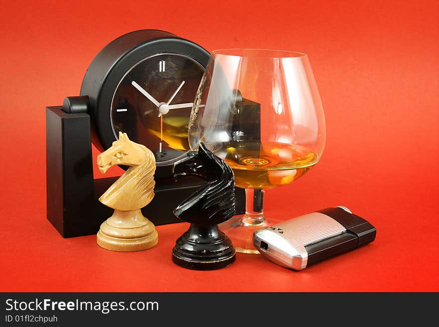 Still life with glass of brandy, chess and clock on red background