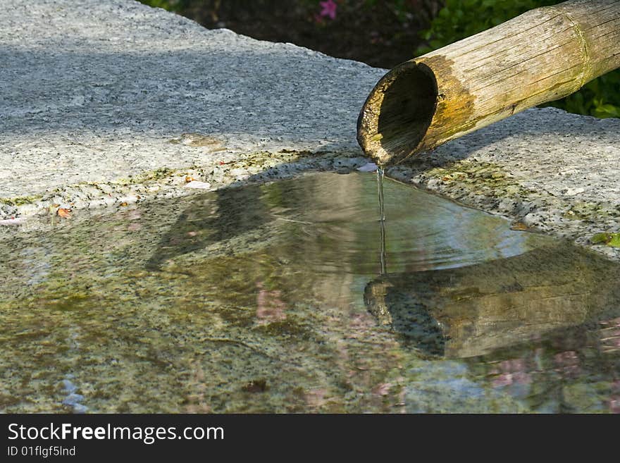 Detail of a Japanese garden. From a single, weathered bamboo spout flows a thin stream of water into a peaceful pool dug out of a large block of rough gray, black, and white granite. The quiet surface of the water in the pool is broken successive rings of ripples from the stream, the pale reflections of blue sky and pink flowers, and a few tiny, fallen leaves and petals. The bamboo shaft casts a l. Detail of a Japanese garden. From a single, weathered bamboo spout flows a thin stream of water into a peaceful pool dug out of a large block of rough gray, black, and white granite. The quiet surface of the water in the pool is broken successive rings of ripples from the stream, the pale reflections of blue sky and pink flowers, and a few tiny, fallen leaves and petals. The bamboo shaft casts a l