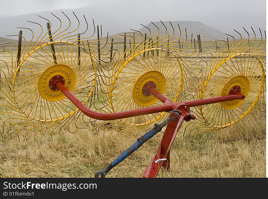 The tines of a large red, yellow, and blue farm implement dominate the foreground of a misty Montana landscape in autumn. Yellow grasses and barbed wire fence stretch out across a cattle ranch toward the mist-enshrouded foothills of the Front Range, near Choteau, Montana. The tines of a large red, yellow, and blue farm implement dominate the foreground of a misty Montana landscape in autumn. Yellow grasses and barbed wire fence stretch out across a cattle ranch toward the mist-enshrouded foothills of the Front Range, near Choteau, Montana.