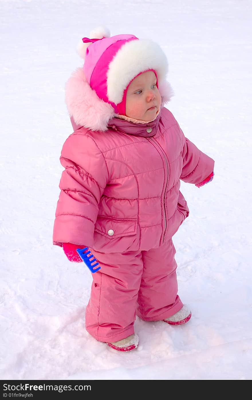 Pretty little girl with blue plastic toy rake in winter outerwear stay on snow-covered ground. Pretty little girl with blue plastic toy rake in winter outerwear stay on snow-covered ground.