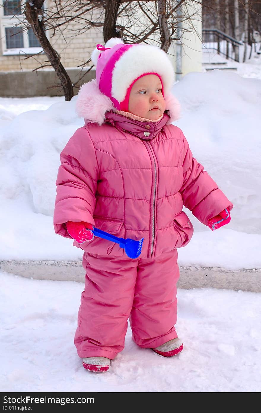 Pretty little girl with blue plastic toy rake in winter outerwear stay on snow-covered ground. Pretty little girl with blue plastic toy rake in winter outerwear stay on snow-covered ground.