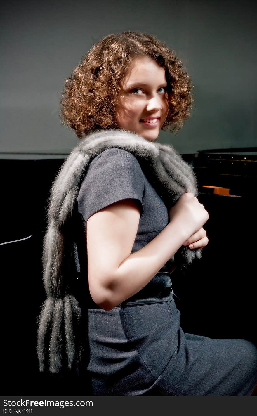 Pretty young dark haired girl with curly hair sitting turned near piano in grey dress with fur scarf smiling isolated