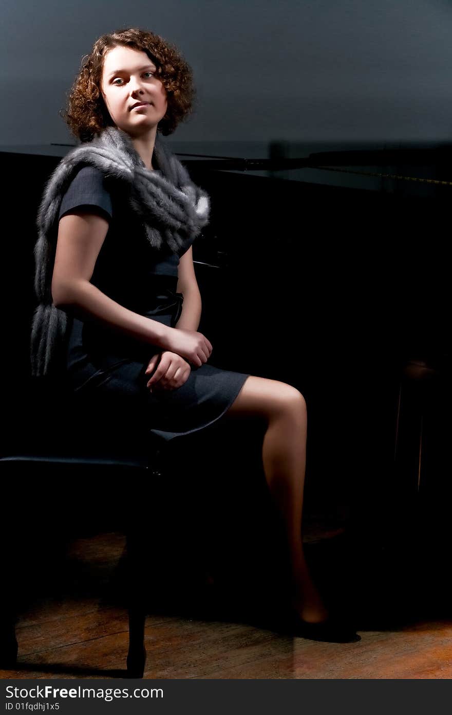 Pretty young lady sitting near black piano in grey dress with fur scarf around neck isolated