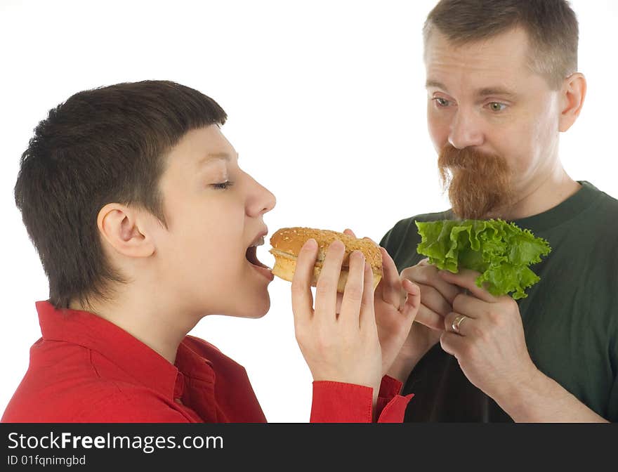 A breaded man with lettuce in hands watching enviously at a woman who is going to bite a hamburger. A breaded man with lettuce in hands watching enviously at a woman who is going to bite a hamburger