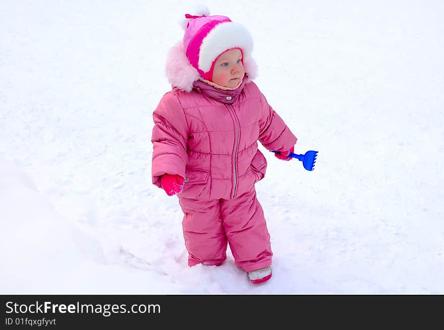 Pretty little girl with blue plastic toy rake in winter outerwear stay on snow-covered ground. Pretty little girl with blue plastic toy rake in winter outerwear stay on snow-covered ground.