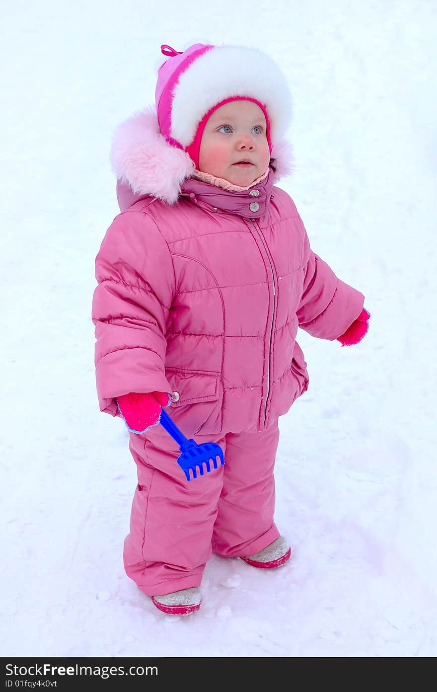 Pretty little girl with blue plastic toy rake in winter outerwear stay on snow-covered ground. Pretty little girl with blue plastic toy rake in winter outerwear stay on snow-covered ground.