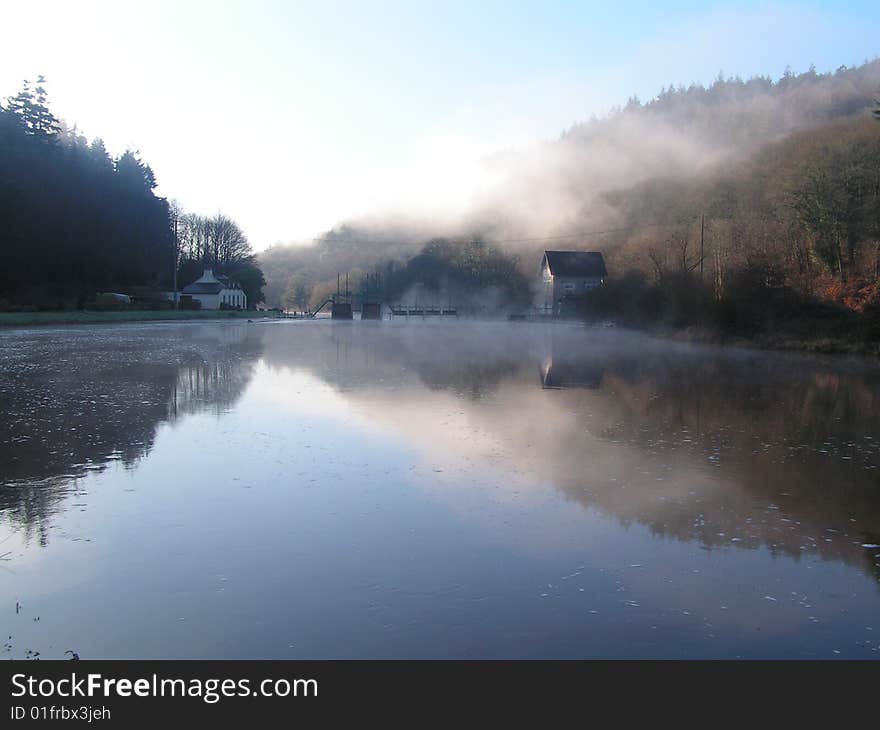 A white building stands beside a still lake whose waters reflect the tree covered hills and mist. A white building stands beside a still lake whose waters reflect the tree covered hills and mist