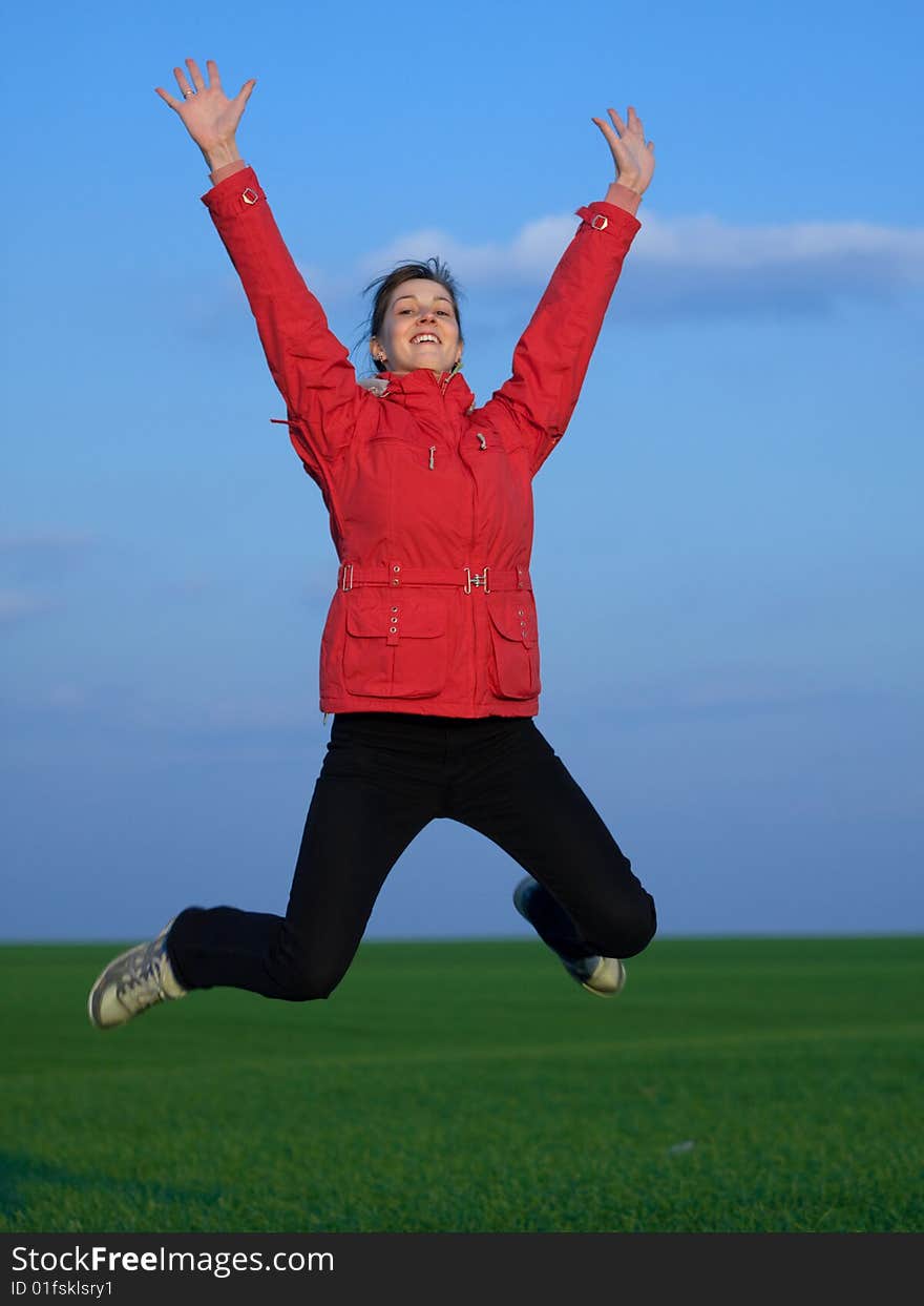 Jumping girl in red jacket on blue sky and green grass background