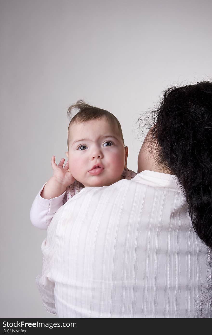 A baby looking over the shoulder of her mom and holding her hand behind her ear like she is asking what you are saying. A baby looking over the shoulder of her mom and holding her hand behind her ear like she is asking what you are saying.