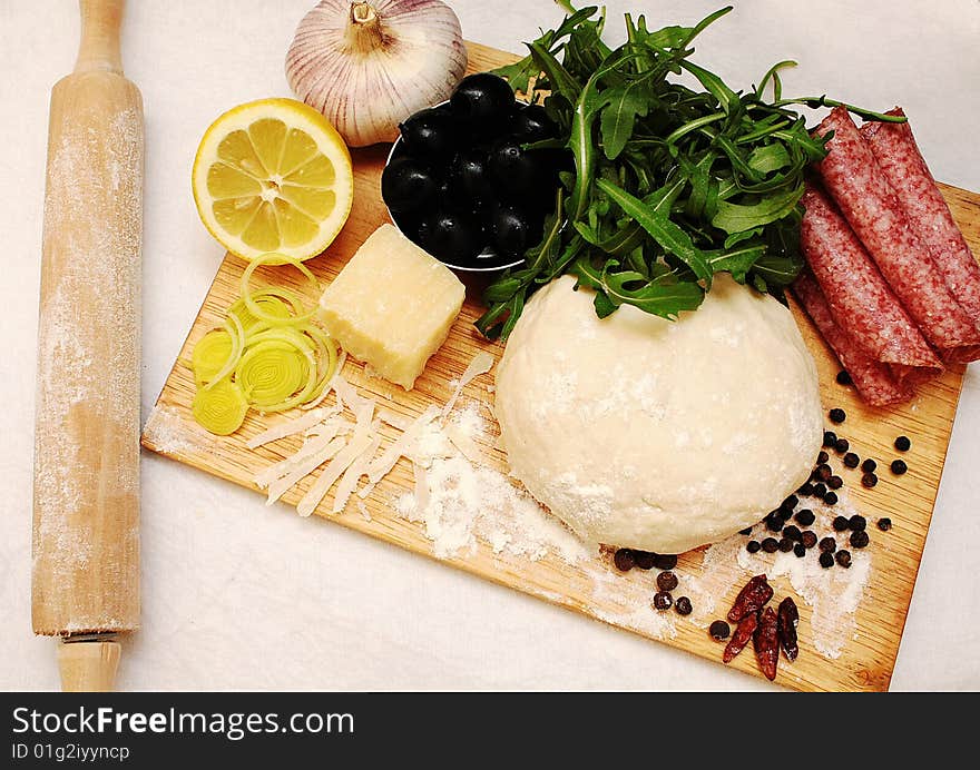 Pizza dough, olives, garlic, rolling pin, rukola, lemon and salami on the board. Pizza dough, olives, garlic, rolling pin, rukola, lemon and salami on the board.