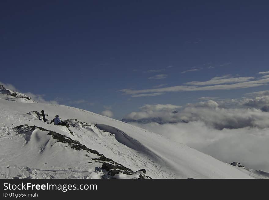 Mountains and low cloud landscape and two figures with thier snowboard sitting down looking at it. Mountains and low cloud landscape and two figures with thier snowboard sitting down looking at it