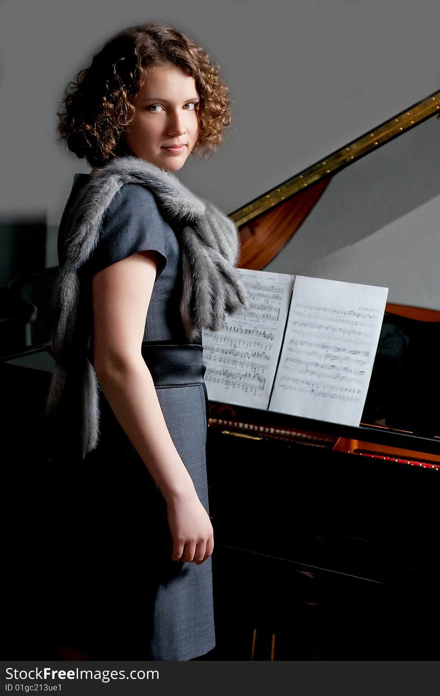 Young girl with serious look in gray dress and fur scarf near black piano with music notes standing turned right looking forward on grey backgorund. Young girl with serious look in gray dress and fur scarf near black piano with music notes standing turned right looking forward on grey backgorund