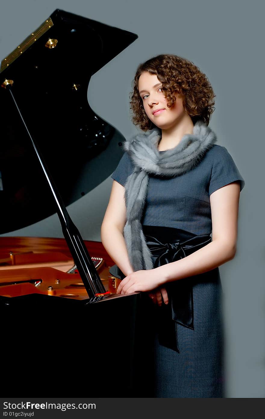 Young girl with curious look in gray dress and fur scarf near black piano  standing turned left with hands crossed appear looking forward on grey backgorund