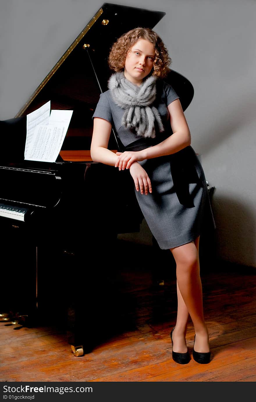 Young girl with curious look in gray dress and fur scarf near black piano standing turned left supported looking forward smiling slaightly on grey backgorund