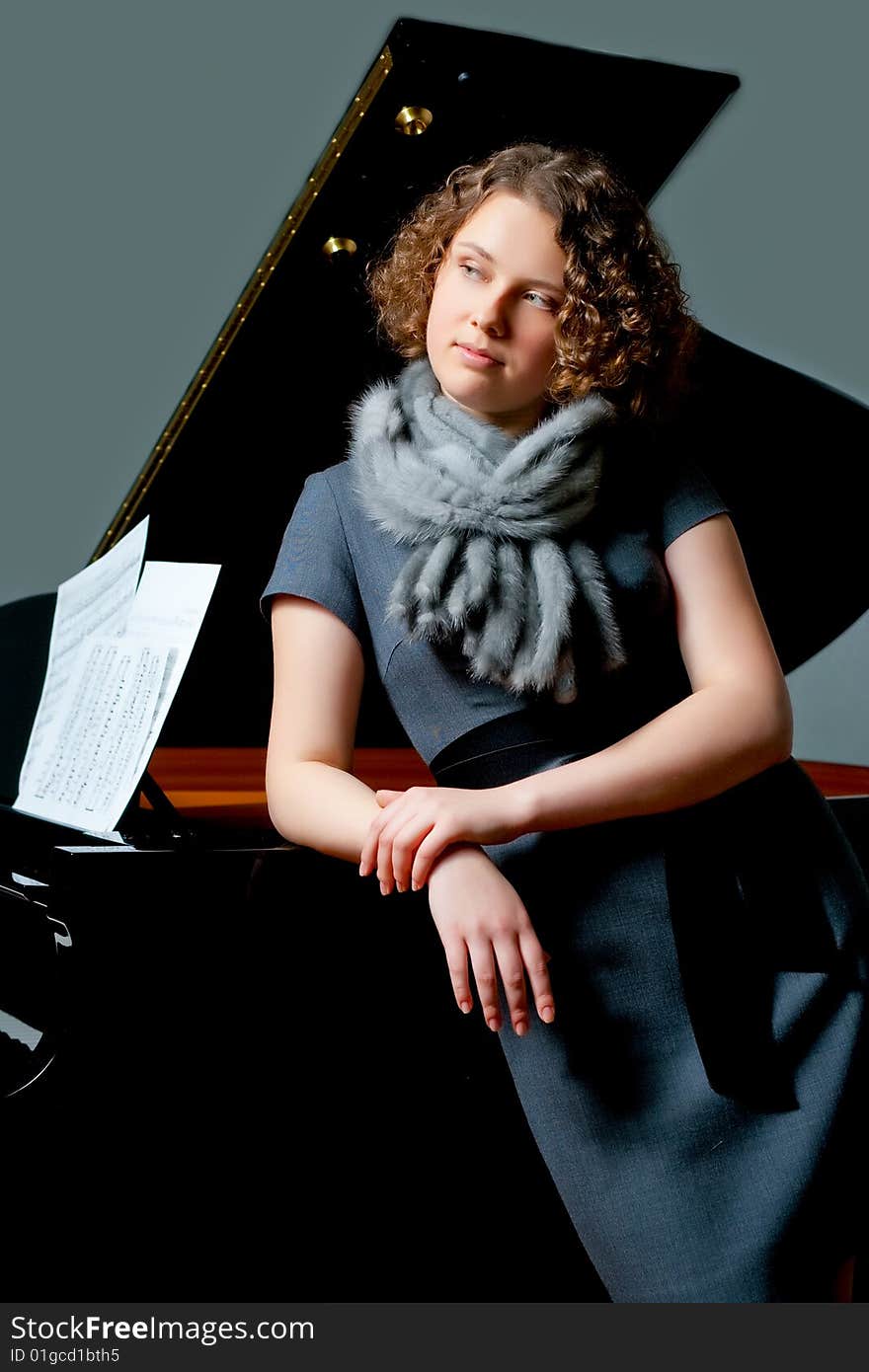 Pretty young girl with curly hair on gray dress and fur scarf standing turned near black piano with hands crossed appear looking forward. Pretty young girl with curly hair on gray dress and fur scarf standing turned near black piano with hands crossed appear looking forward