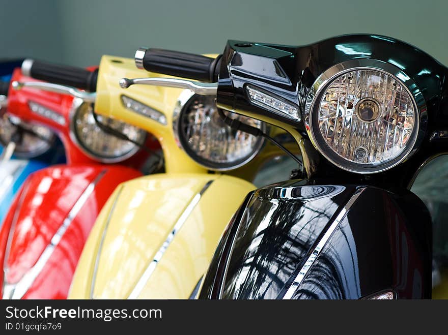 Headlights of three scooters, black, yellow and red. Shallow depth of field with the first scooter in focus.