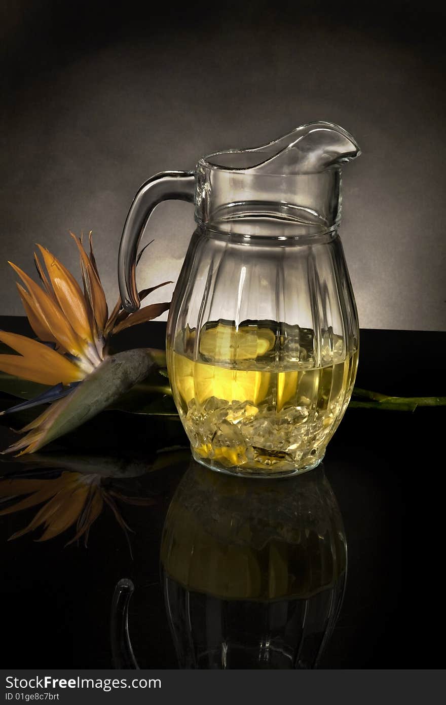 Glass pitcher filled with lemonade, next to a bird of paradise flower on a black reflective surface, against a gray background with a spot light. Glass pitcher filled with lemonade, next to a bird of paradise flower on a black reflective surface, against a gray background with a spot light.