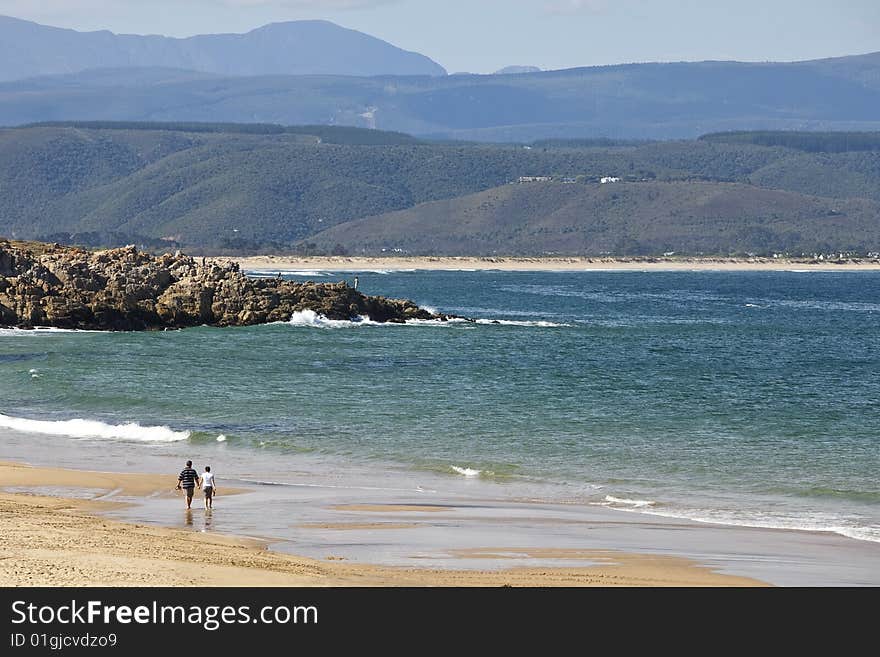 A middle-aged couple strolls along the beach with mountains in the distance and bright blue ocean. A middle-aged couple strolls along the beach with mountains in the distance and bright blue ocean.