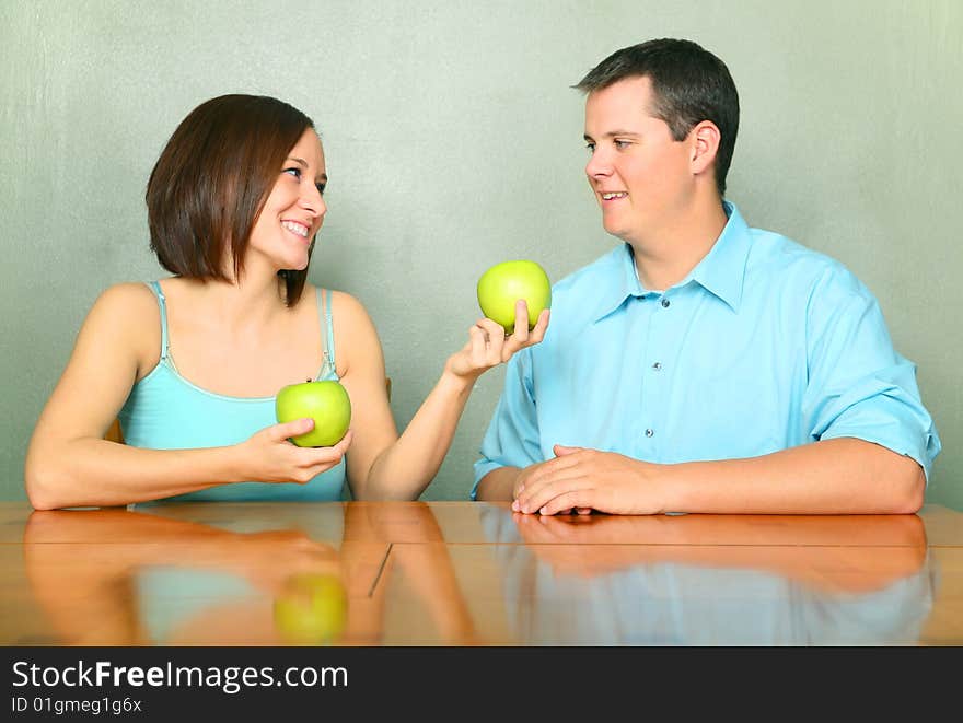 Beautiful young female caucasian offering green apple to her boyfriend or husband on kitchen table. Beautiful young female caucasian offering green apple to her boyfriend or husband on kitchen table