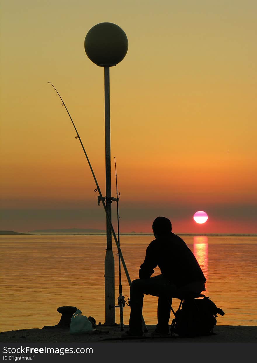 A fisherman’s silhouette sitting in a pier by the sunset. A fisherman’s silhouette sitting in a pier by the sunset