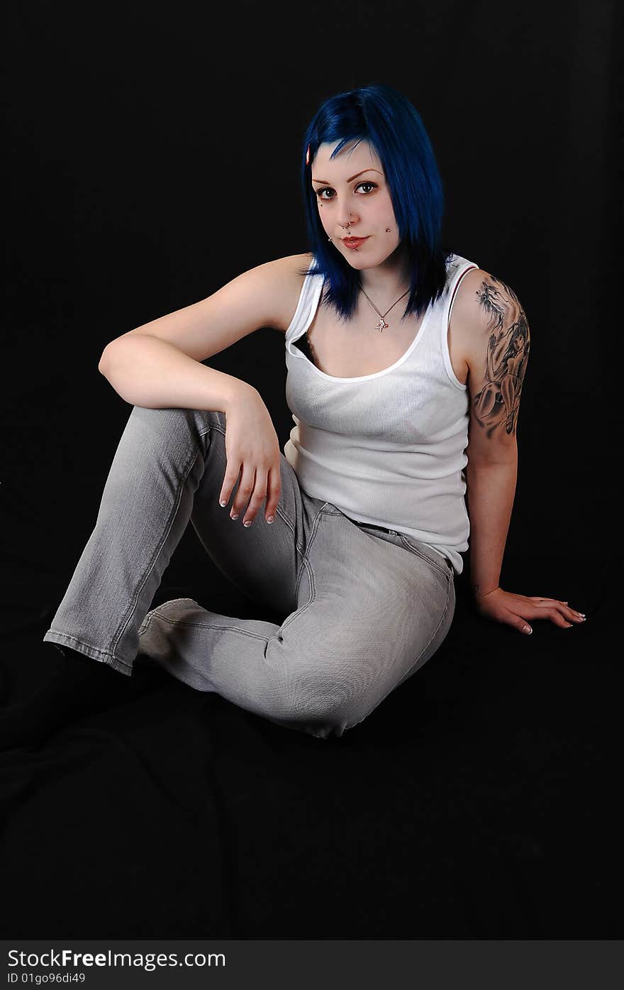 Pretty young blue haired girl in white t-shirt and jeans with a big tattoo on her arm sitting on the floor for black background. Pretty young blue haired girl in white t-shirt and jeans with a big tattoo on her arm sitting on the floor for black background.