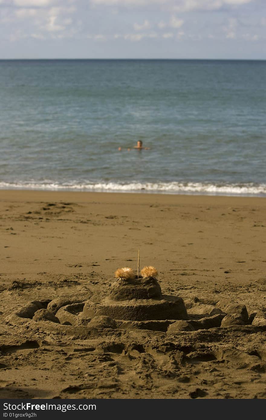 A sand castle in the forground and a soft focus person floating in the ocean in the background