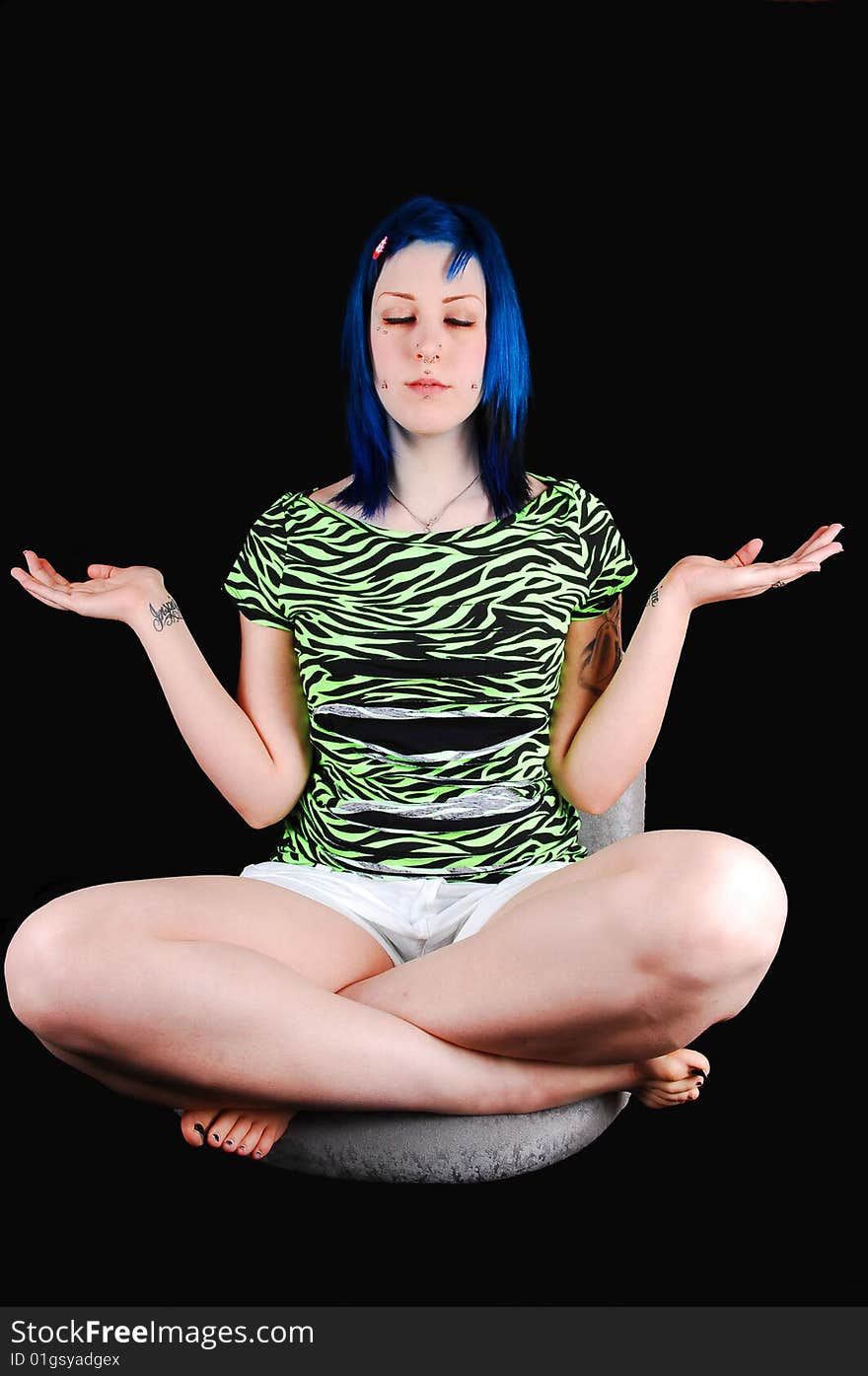 Pretty young blue haired girl is meditating in a green top and white 
shorts, sitting on a chair for black background. Pretty young blue haired girl is meditating in a green top and white 
shorts, sitting on a chair for black background.