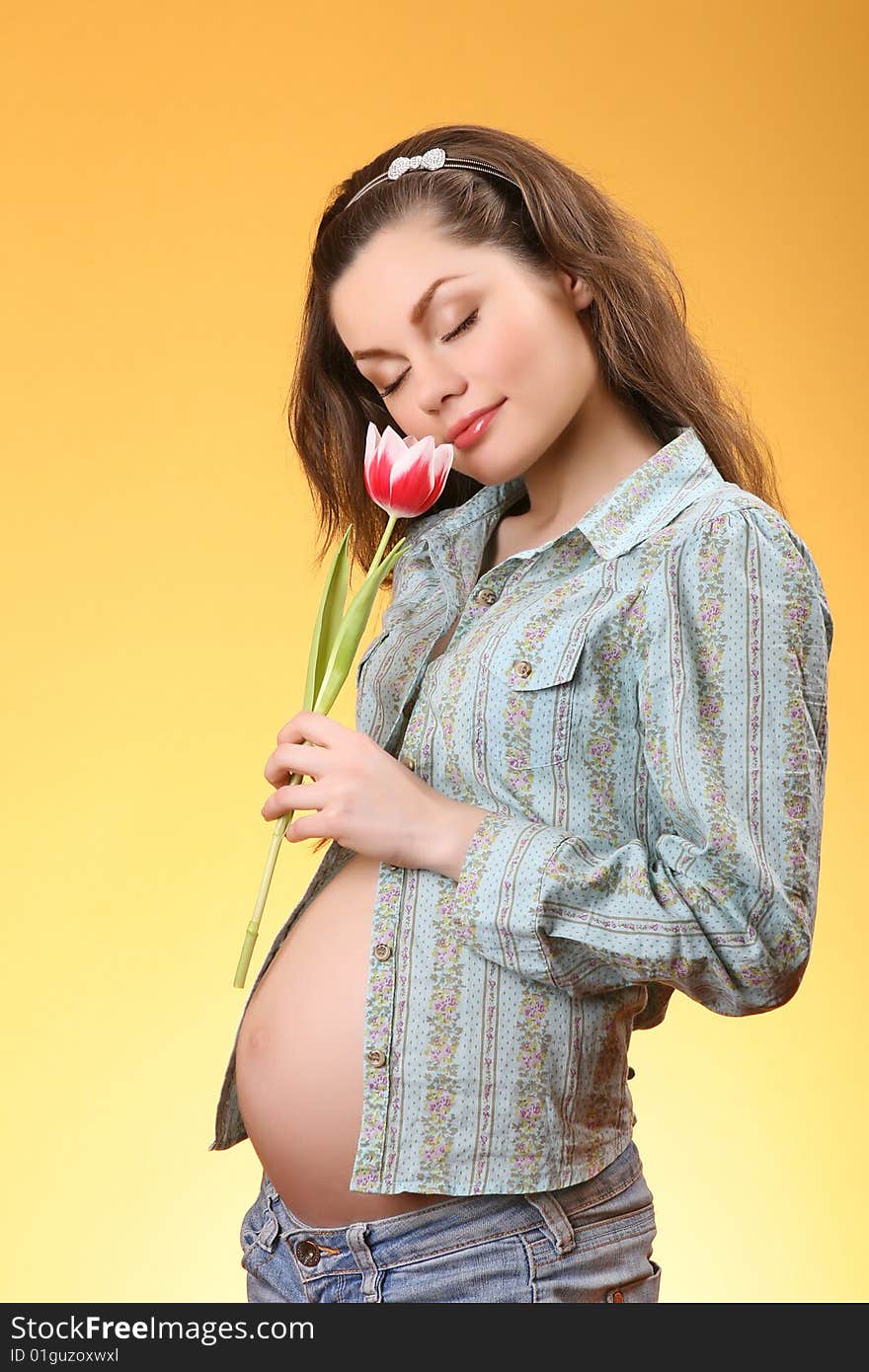 Young pregnant woman holding a tulip with eyes closed on yellow background