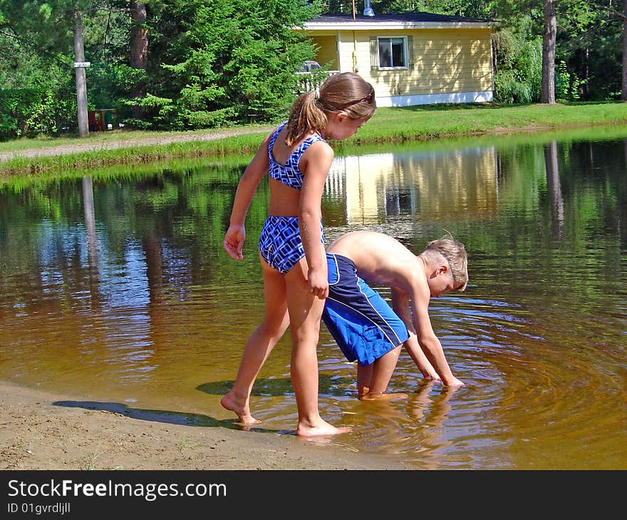 A boy is trying to catch some small fish in the lake while a girl looks on. A boy is trying to catch some small fish in the lake while a girl looks on