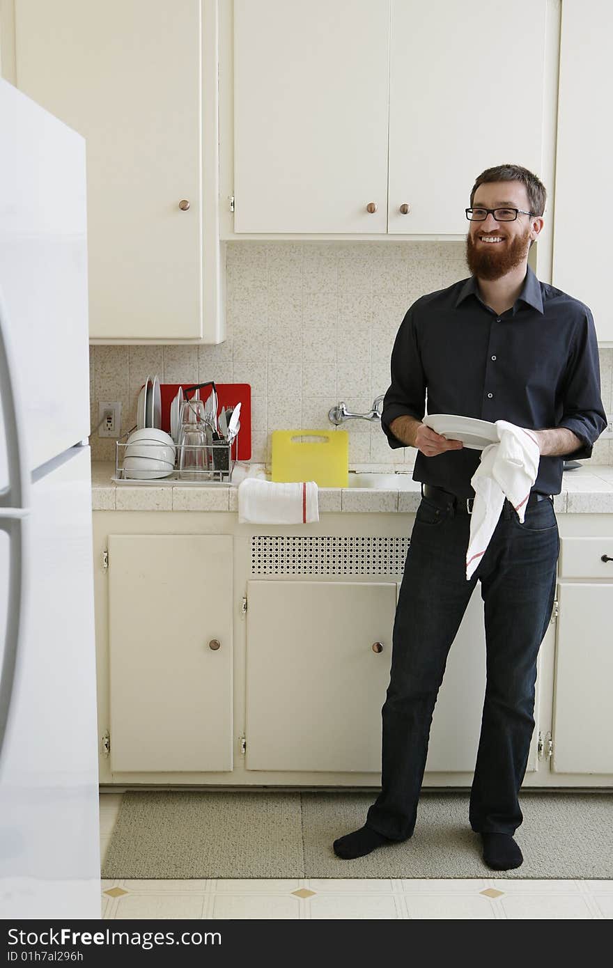 Man with beard smiles while helping in the kitchen.  He is wearing a dark shirt and denim is standing by sink. Man with beard smiles while helping in the kitchen.  He is wearing a dark shirt and denim is standing by sink.