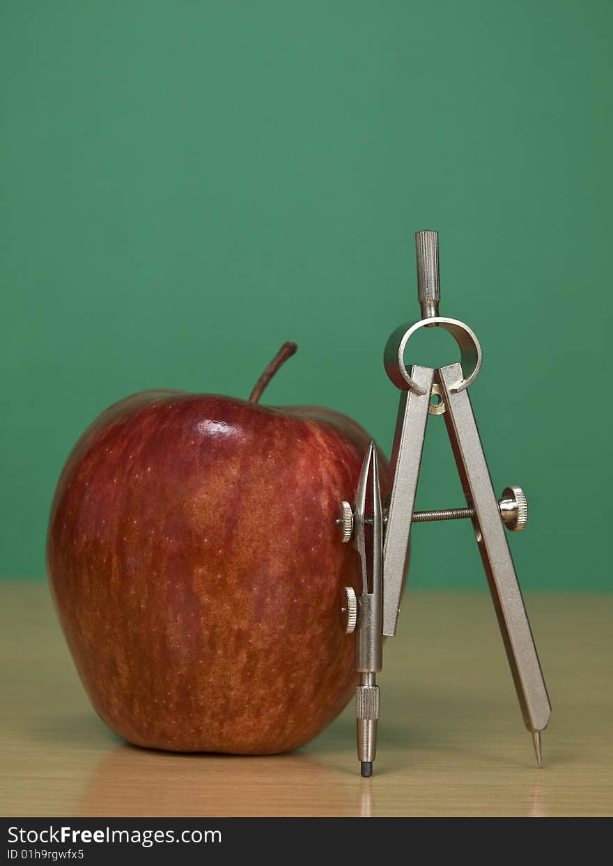 A red apple and a drawing compass over a classroom desk. Blank chalkboard on the background. A red apple and a drawing compass over a classroom desk. Blank chalkboard on the background.