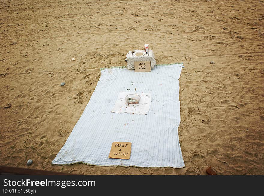 A panhandlers blanket, on the beach, with a dinner palcesetting, set up to accept donations. A panhandlers blanket, on the beach, with a dinner palcesetting, set up to accept donations.