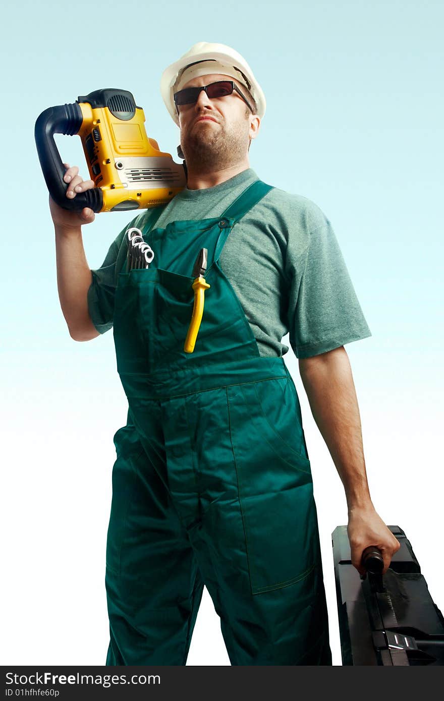 Serious skeptic workman in helmet, black glasses and overalls hold perforator on the shoulder and big black suitcase over white background. Serious skeptic workman in helmet, black glasses and overalls hold perforator on the shoulder and big black suitcase over white background